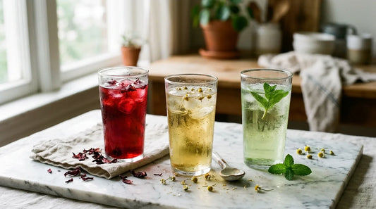 Three glasses of iced caffeine-free herbal tea — hibiscus, chamomile, and peppermint — on a marble surface with dried petals and fresh mint