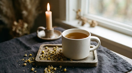 White ceramic mug of golden chamomile tea surrounded by dried chamomile flowers on dark linen with soft candlelight for a cozy evening