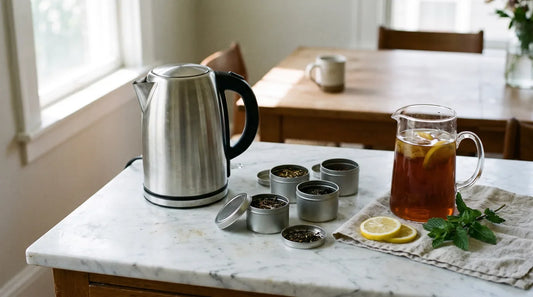 Electric kettle on marble countertop with loose leaf tea tins, amber iced tea pitcher, lemon wedges, and fresh mint for a Labor Day tea station