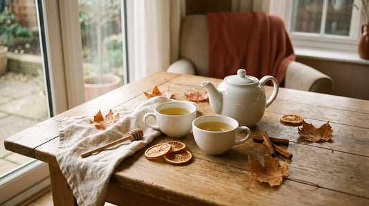Fall brunch tea spread with a white ceramic teapot, golden green tea cups, cinnamon sticks, dried orange slices, and amber autumn leaves on a weathered oak table