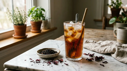 Tall glass of deep amber iced tea with clear ice cubes on a marble surface surrounded by dried hibiscus petals and loose black tea leaves