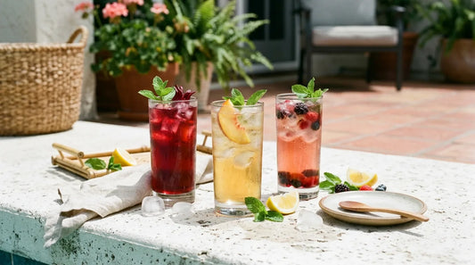 Three tall glasses of poolside iced tea — ruby hibiscus, golden peach, and blush berry — on a white concrete surface with ice cubes, lemon wedges, and mint sprigs in summer light