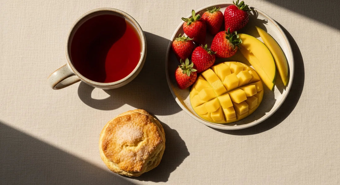 Ceramic cup of hibiscus tea beside fresh strawberries, mango slices, and a butter scone on a linen surface in spring light