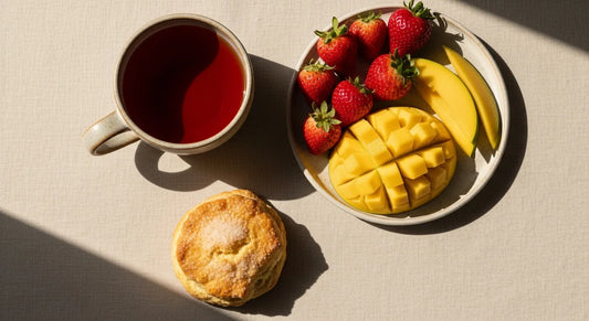 Ceramic cup of hibiscus tea beside fresh strawberries, mango slices, and a butter scone on a linen surface in spring light
